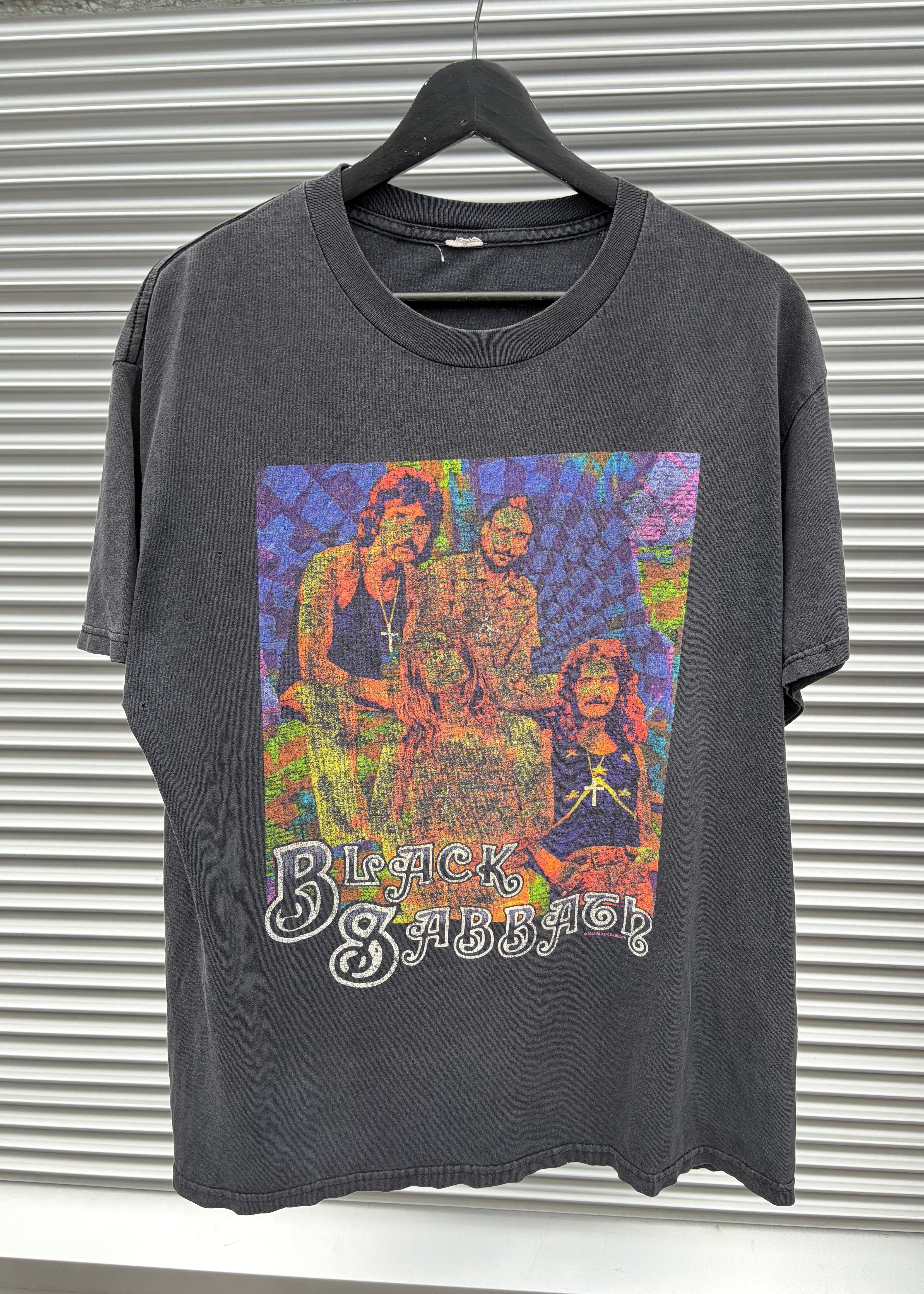 A faded black graphic t-shirt featuring the band Black Sabbath, laid flat on a gray corrugated metal background. The shirt has a large, colorful, psychedelic-style print of the band members on the front with the band's name in white gothic lettering.